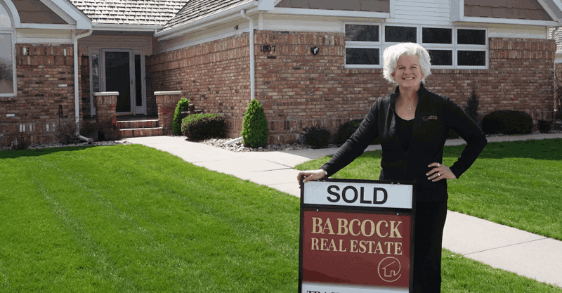 Tracy in front of a house standing by a sold sign.